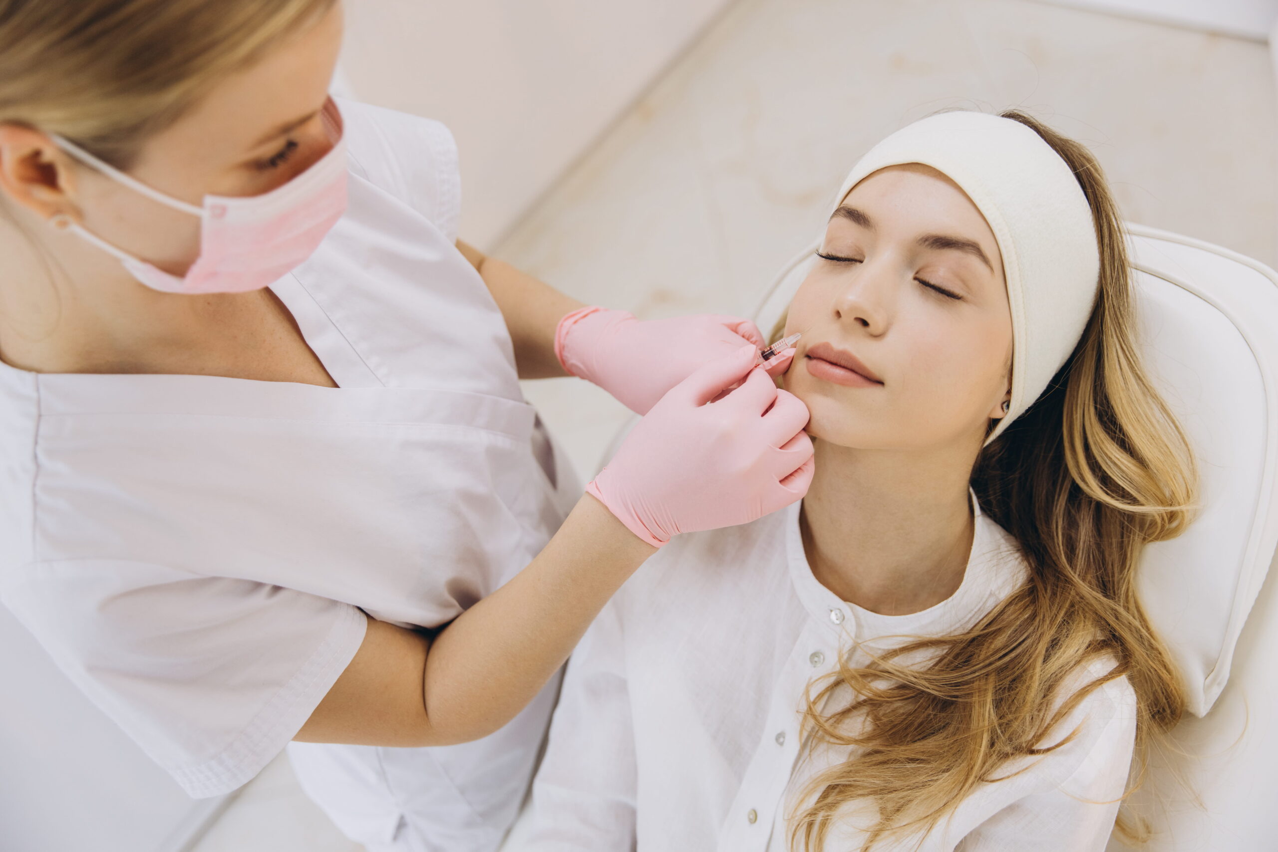 Cosmetologist making botox injection into lips of young blond woman lying on couch in beauty salon